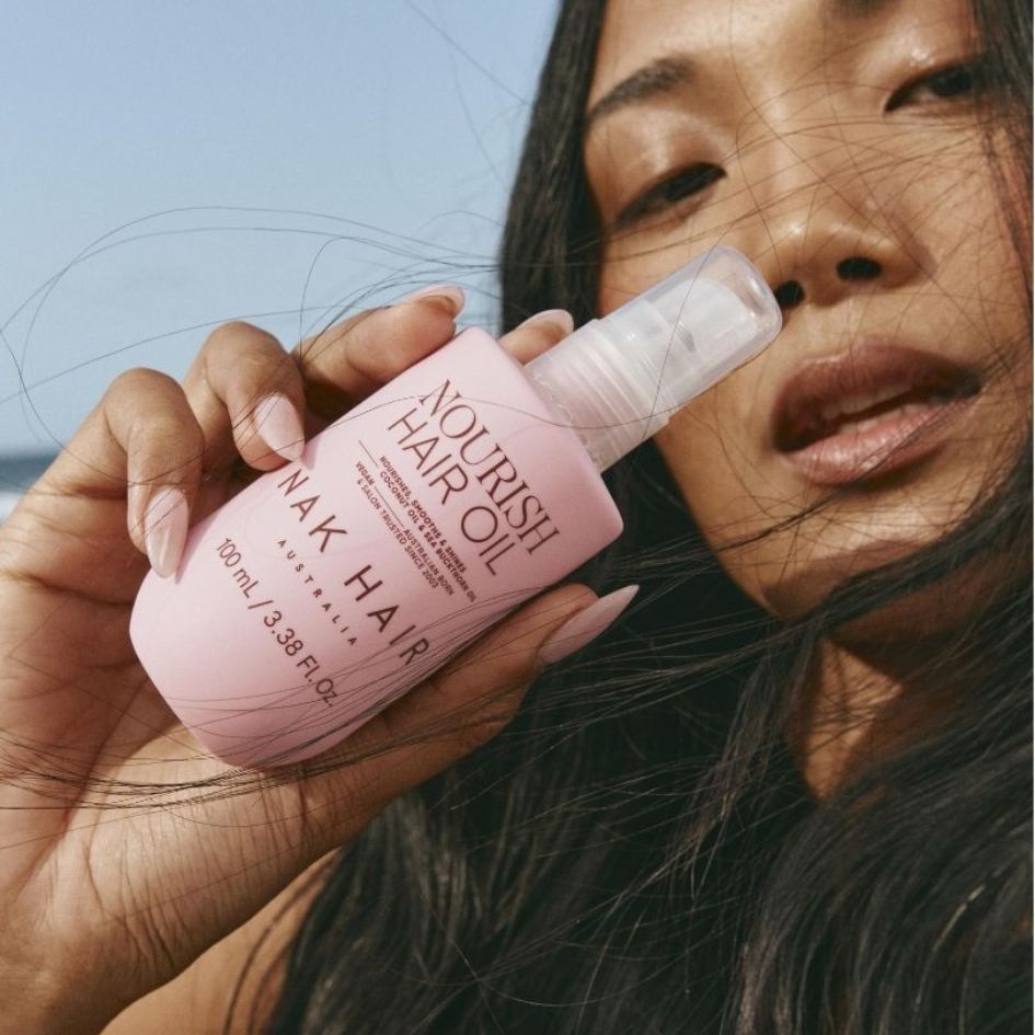 NEW NAK Signature Nourish Hair Oil Vrouw houdt een roze fles NAK Hair Nourish Hair Oil vast, met de tekst ‘Nourish Hair Oil’ op het etiket